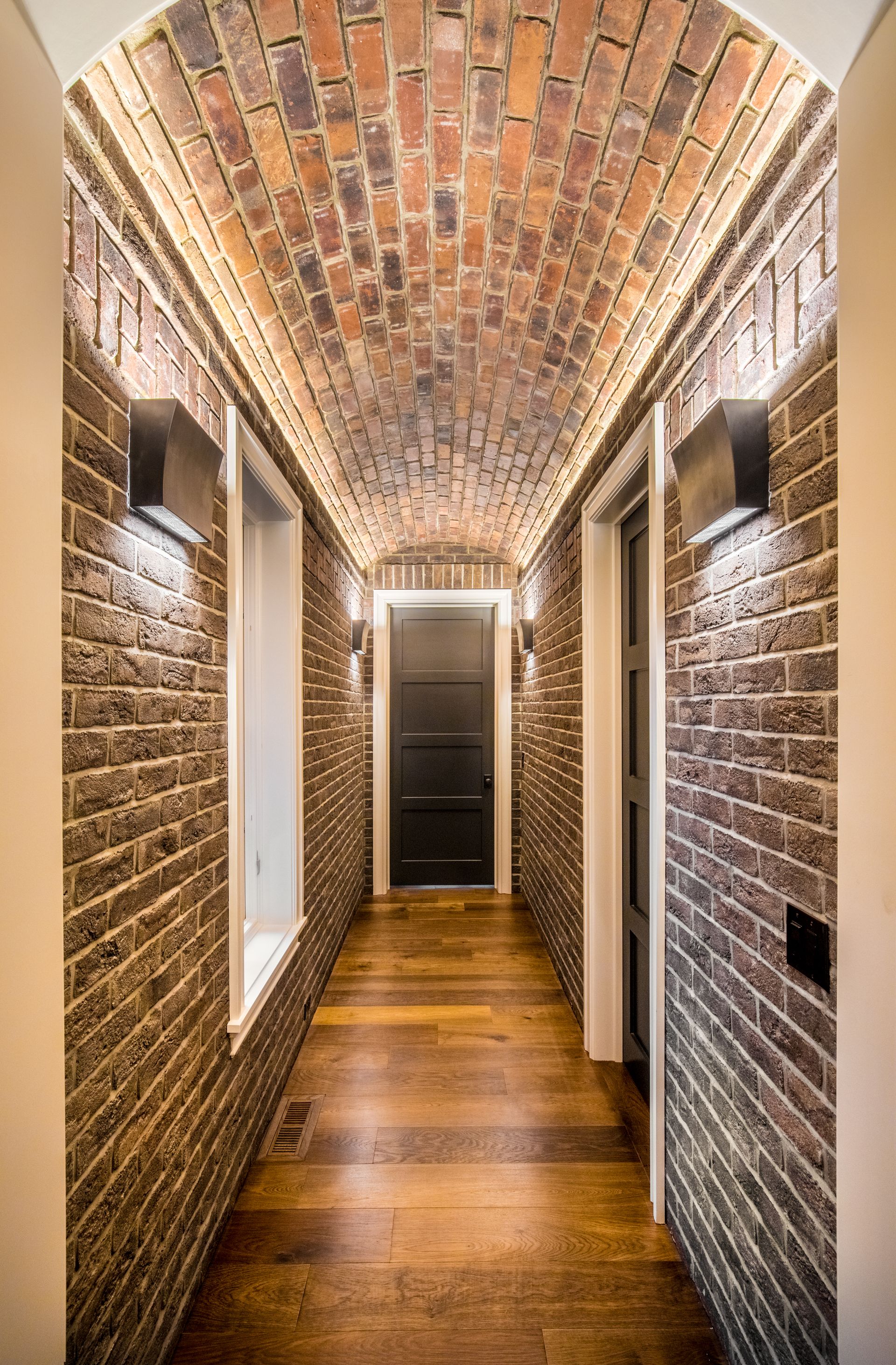 Narrow hallway with brick walls and arched ceiling, wood floor, and black door.