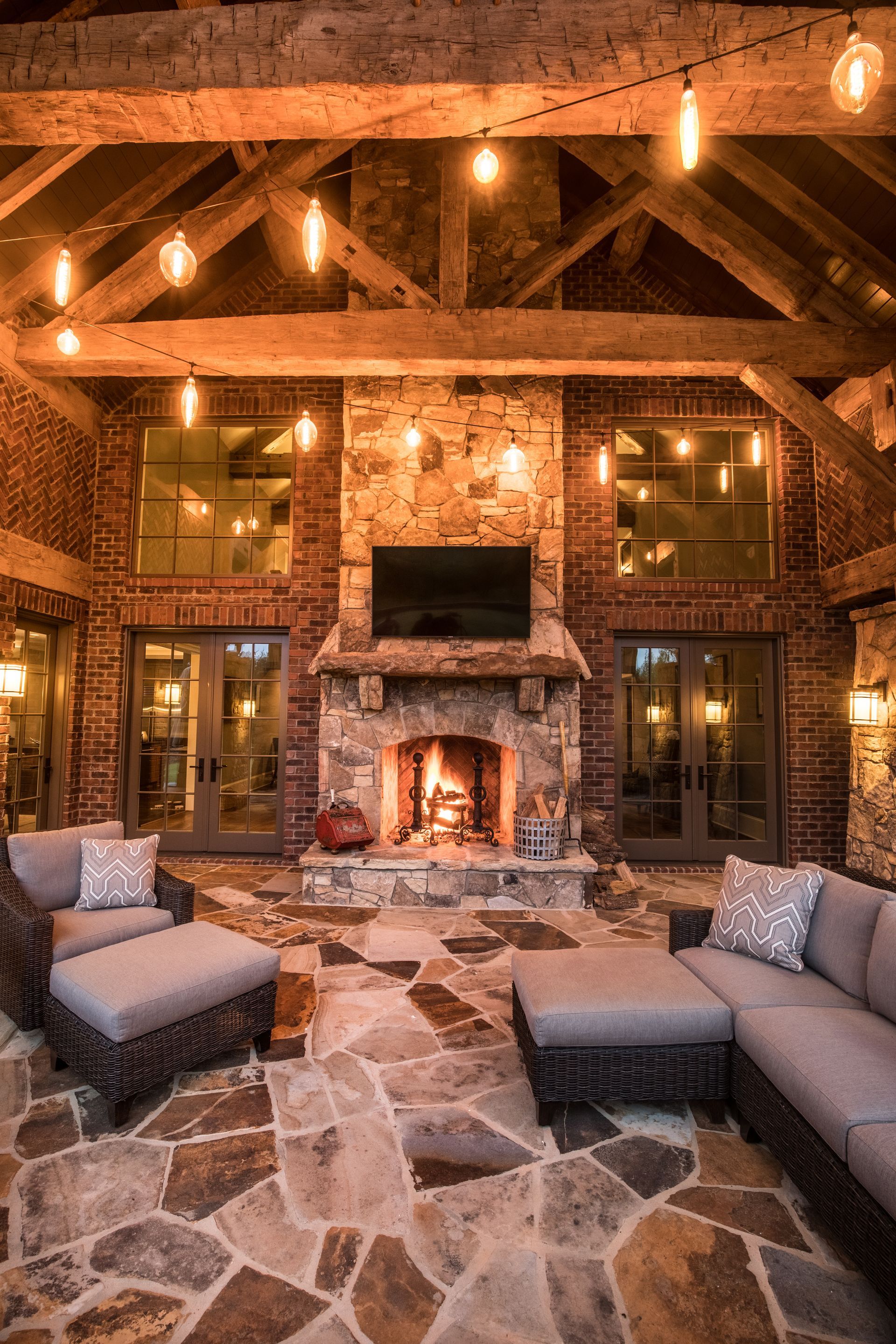 Cozy outdoor patio with stone fireplace, comfortable seating, string lights, and exposed wooden beams.