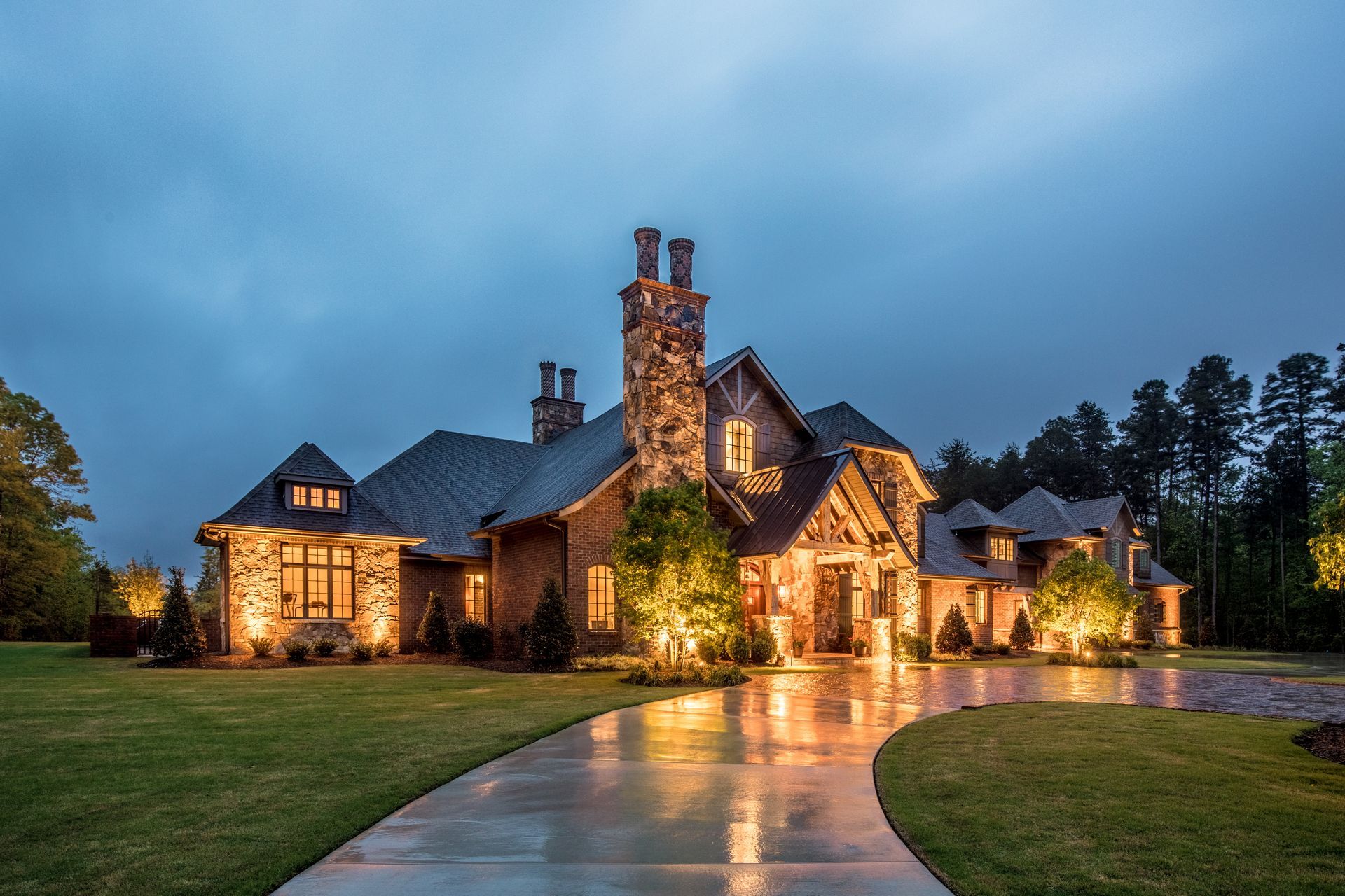Luxury stone house with illuminated driveway and exterior lights at dusk.
