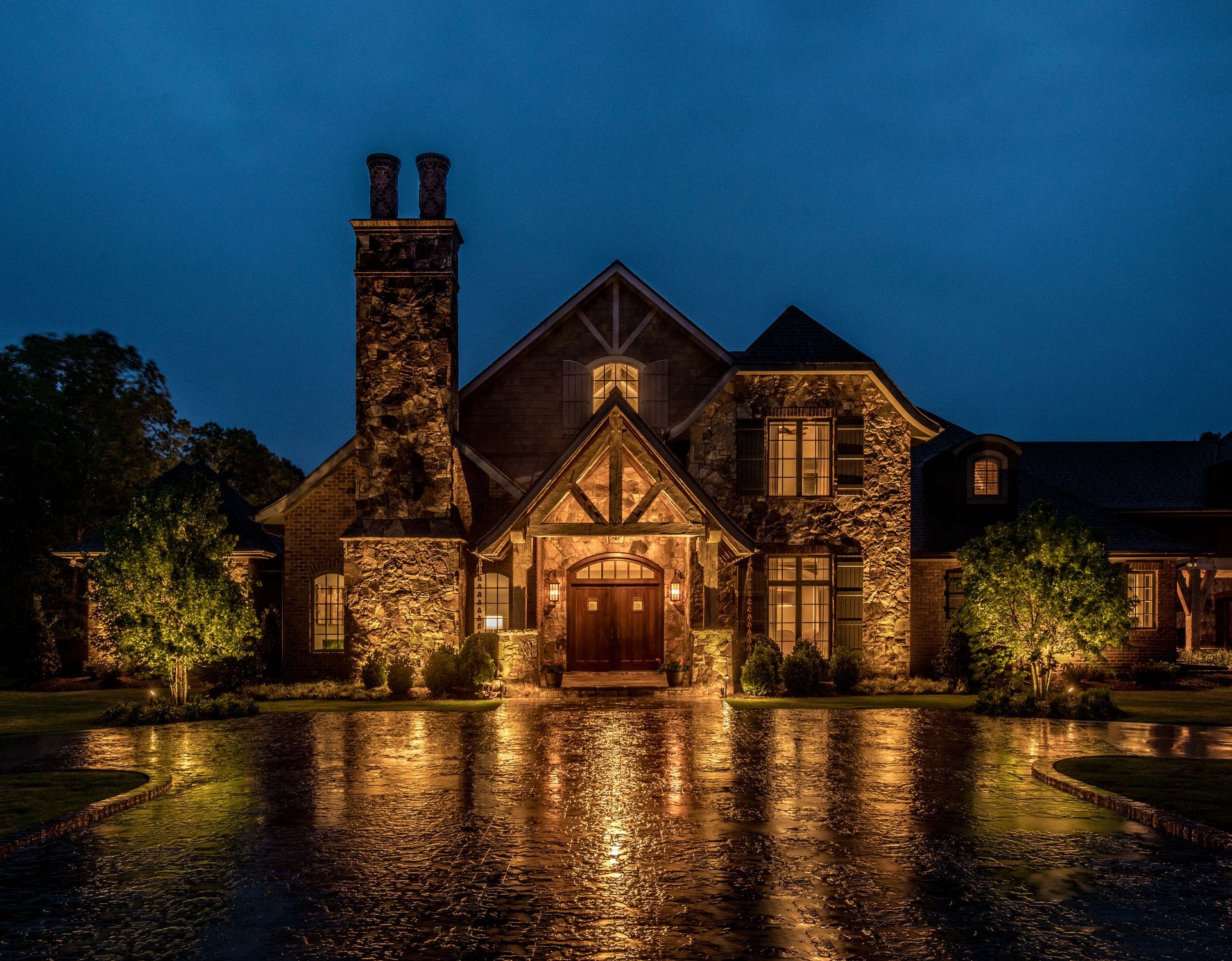 Stone house lit up at night, reflecting in a wet driveway. Dark blue sky overhead.