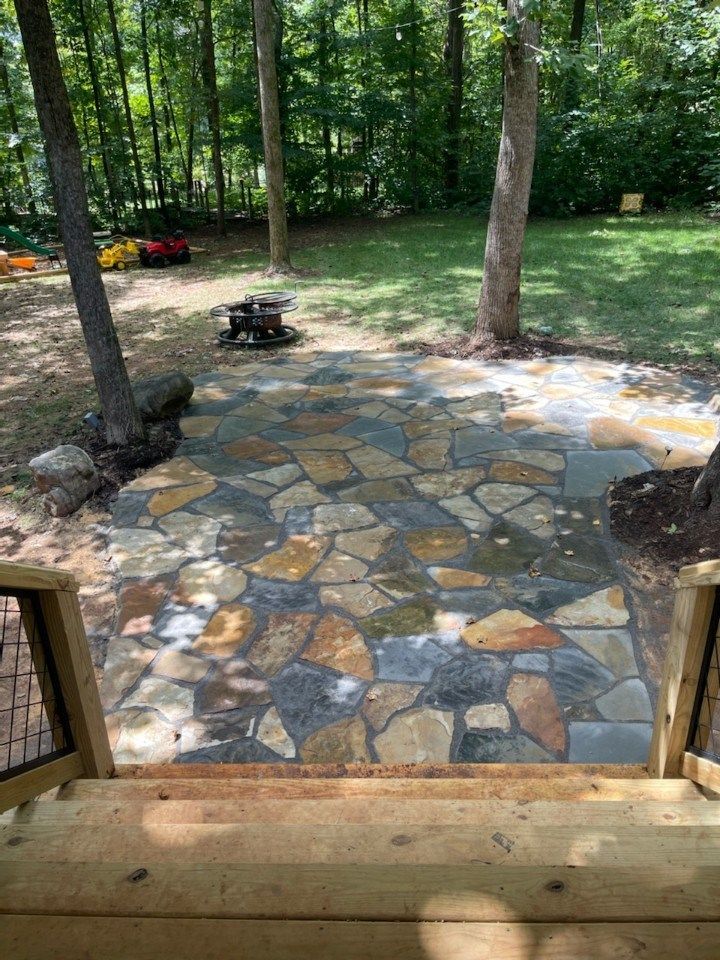 A stone patio made of irregular, multicolored flagstones viewed from the bottom of wooden steps in a wooded backyard.