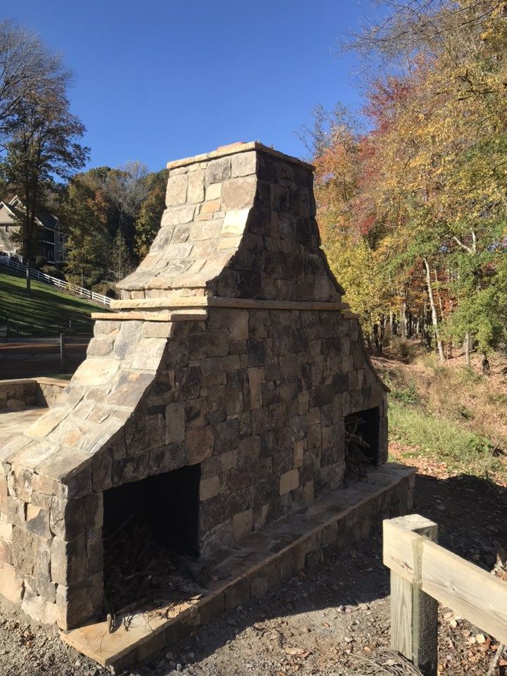 A double-sided stone outdoor fireplace stands in a grassy park setting near trees with autumn foliage.