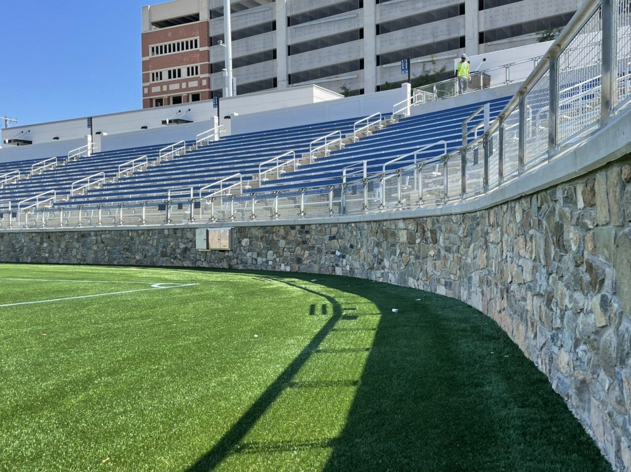 A curved stadium wall of stone surrounds a green artificial turf field and blue stadium seating.