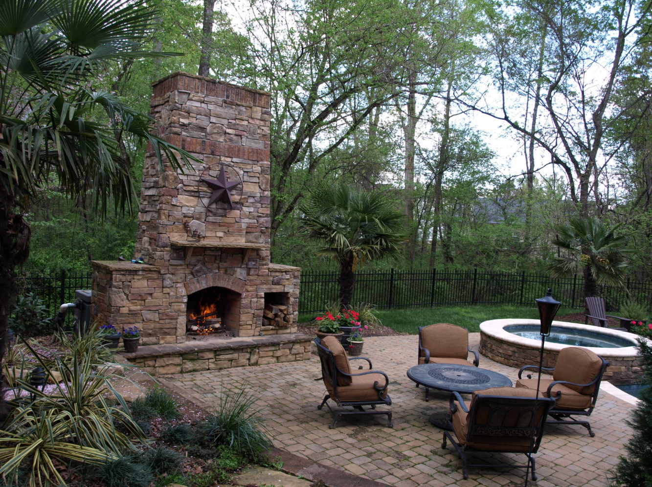 Stone outdoor fireplace with seating area, adjacent to a spa, in a lush backyard.
