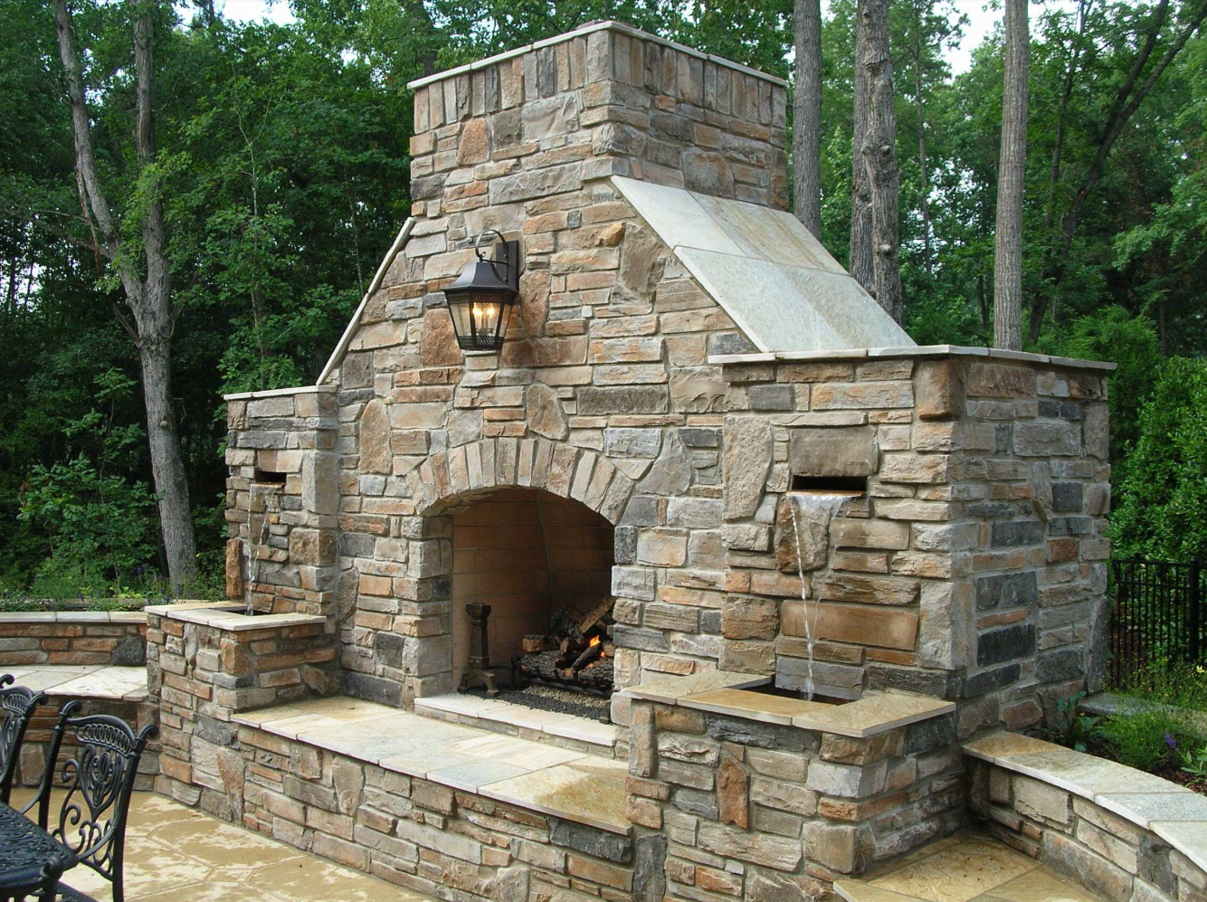 Stone outdoor fireplace with lit interior, in a backyard setting.