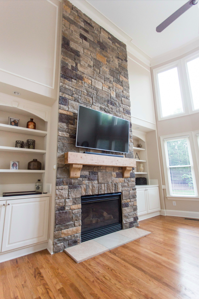 Stone fireplace with built-in shelves and a mounted TV. Wooden mantel, hardwood floor, and large windows.