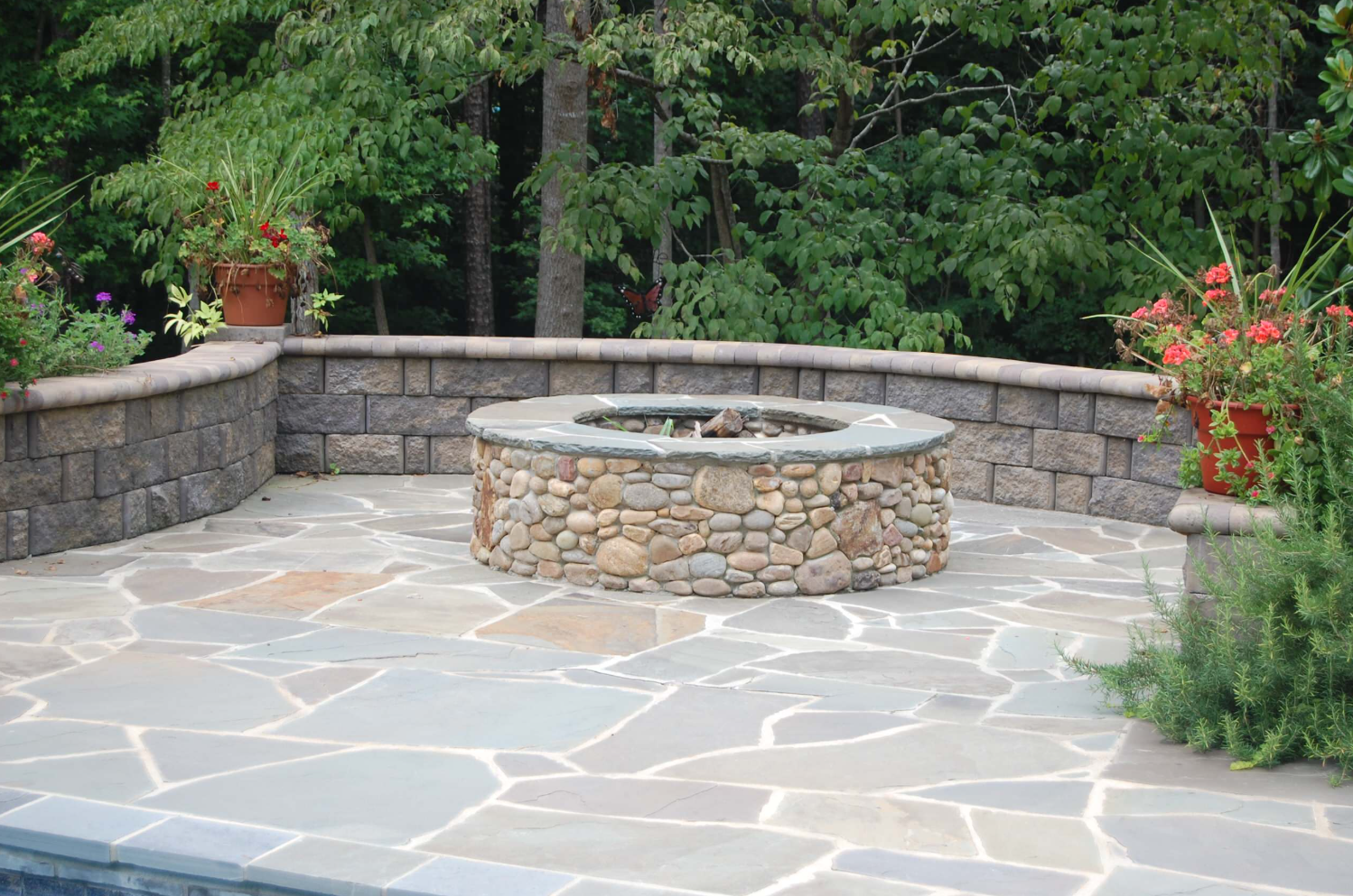 Stone fire pit on a flagstone patio, surrounded by stone walls and potted plants, wooded background.