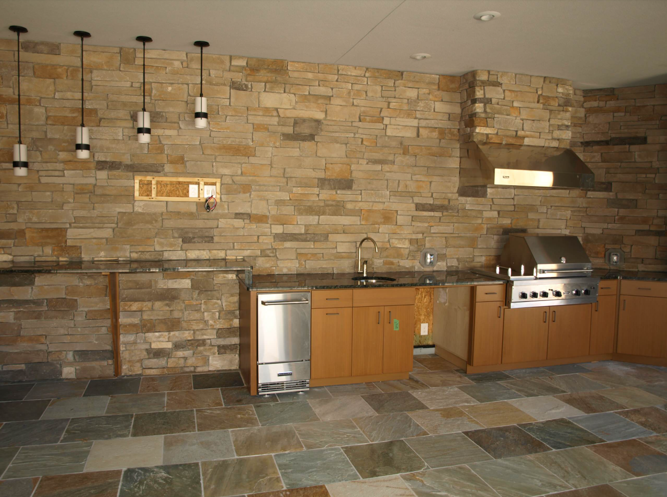 Outdoor kitchen with stone wall, wooden cabinets, stainless steel appliances, and slate floor.
