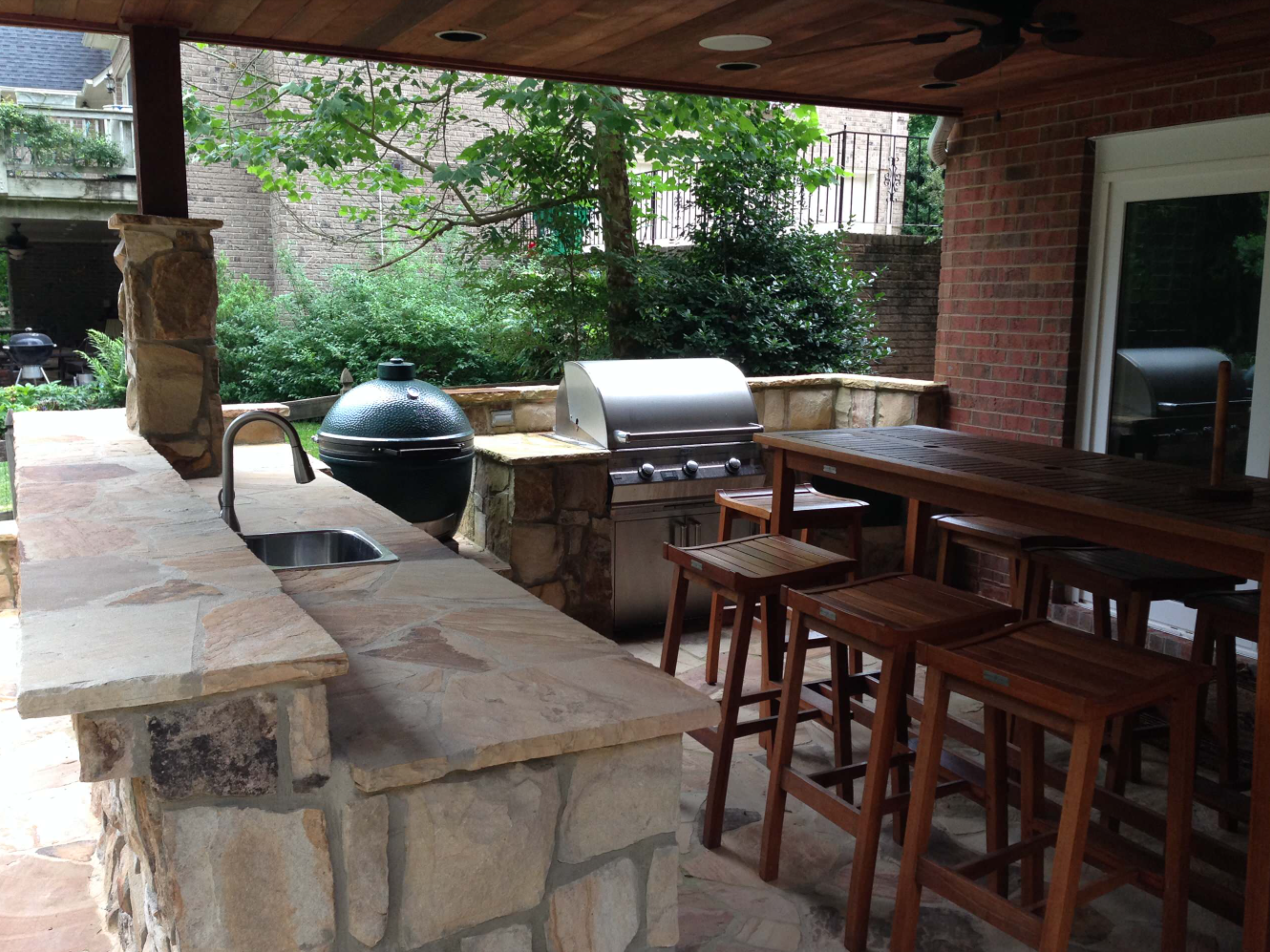 Outdoor kitchen with stone counters, grill, sink, Big Green Egg, and bar with stools.