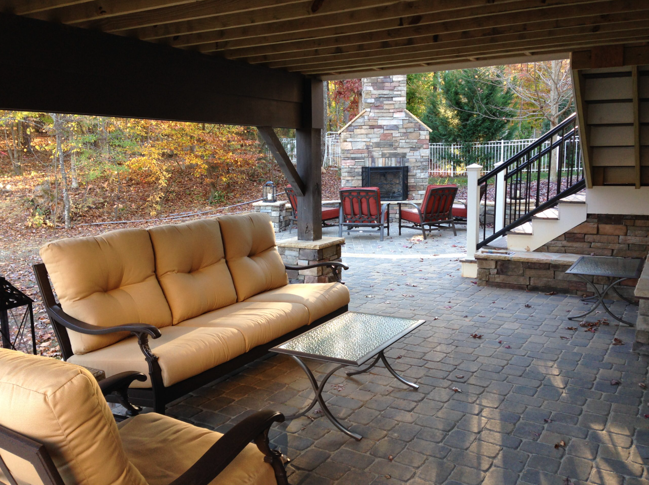 Patio with cushioned furniture under a wooden deck, stone fireplace in the background. Autumn leaves visible.
