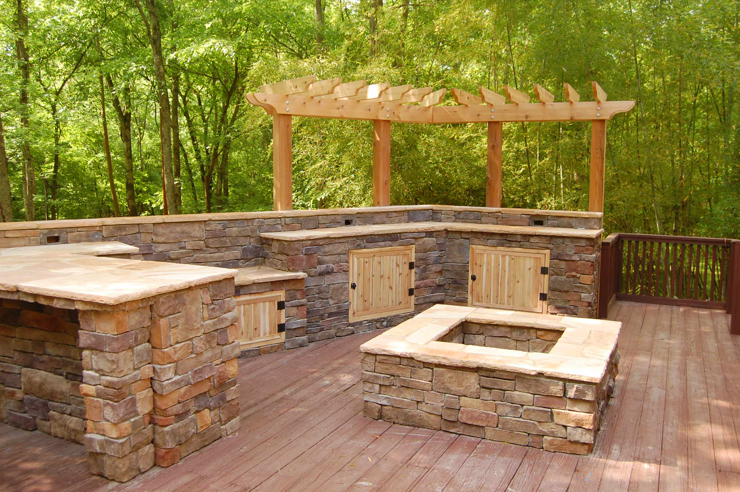 Outdoor kitchen with stone counters, wooden pergola, and fire pit on a wooden deck surrounded by trees.