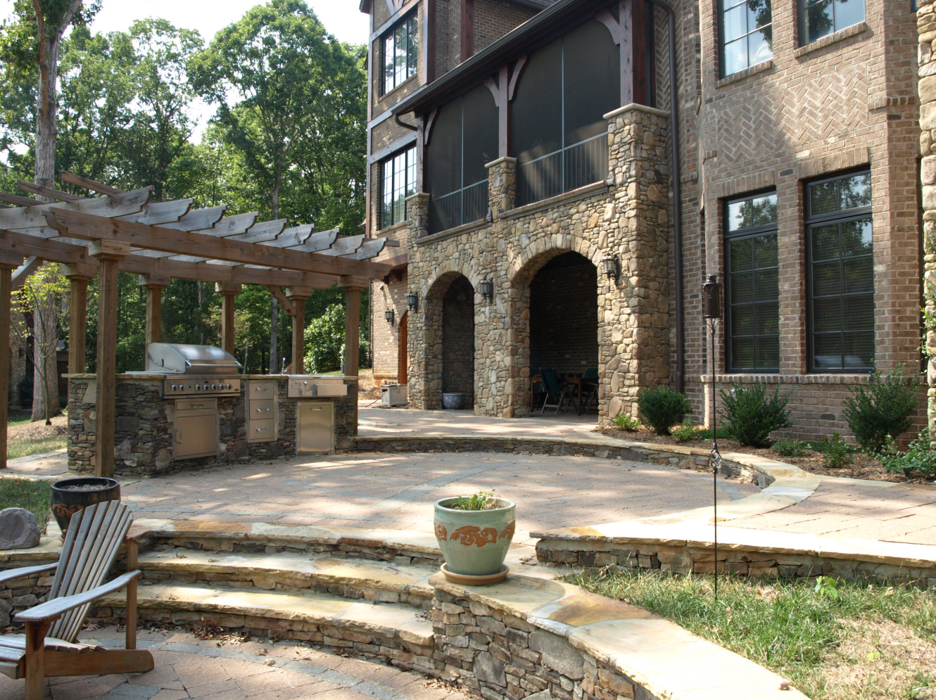 Stone patio with outdoor kitchen, pergola, and brick house.