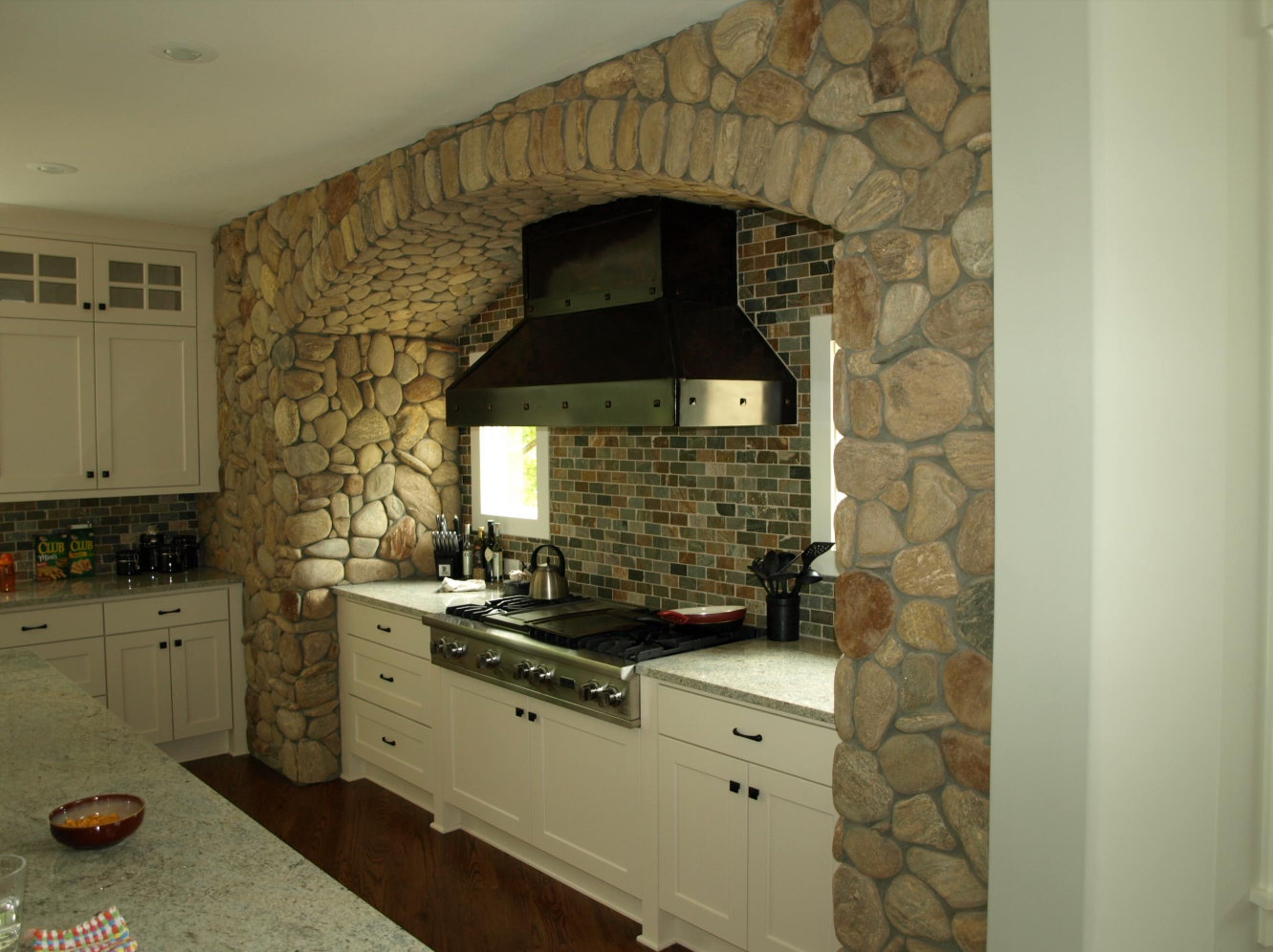Kitchen with stone wall and archway, white cabinets, range hood, and stove.