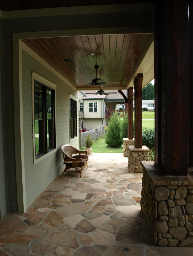 Covered porch with flagstone floor, wood ceiling, stone columns, wicker chair, and view of a yard.