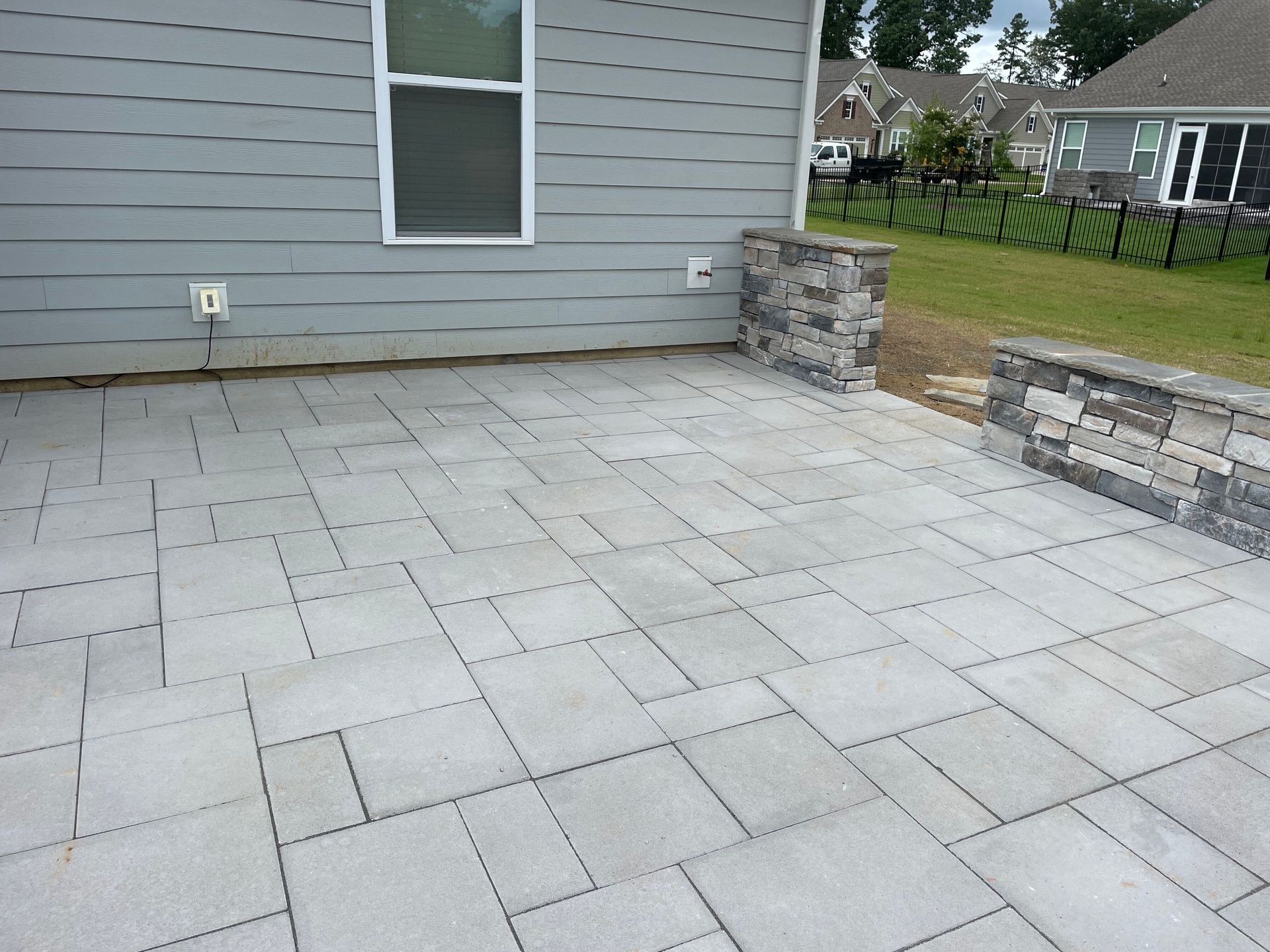 Gray paver patio with stacked stone pillars against a gray house, next to a grassy yard and fence.