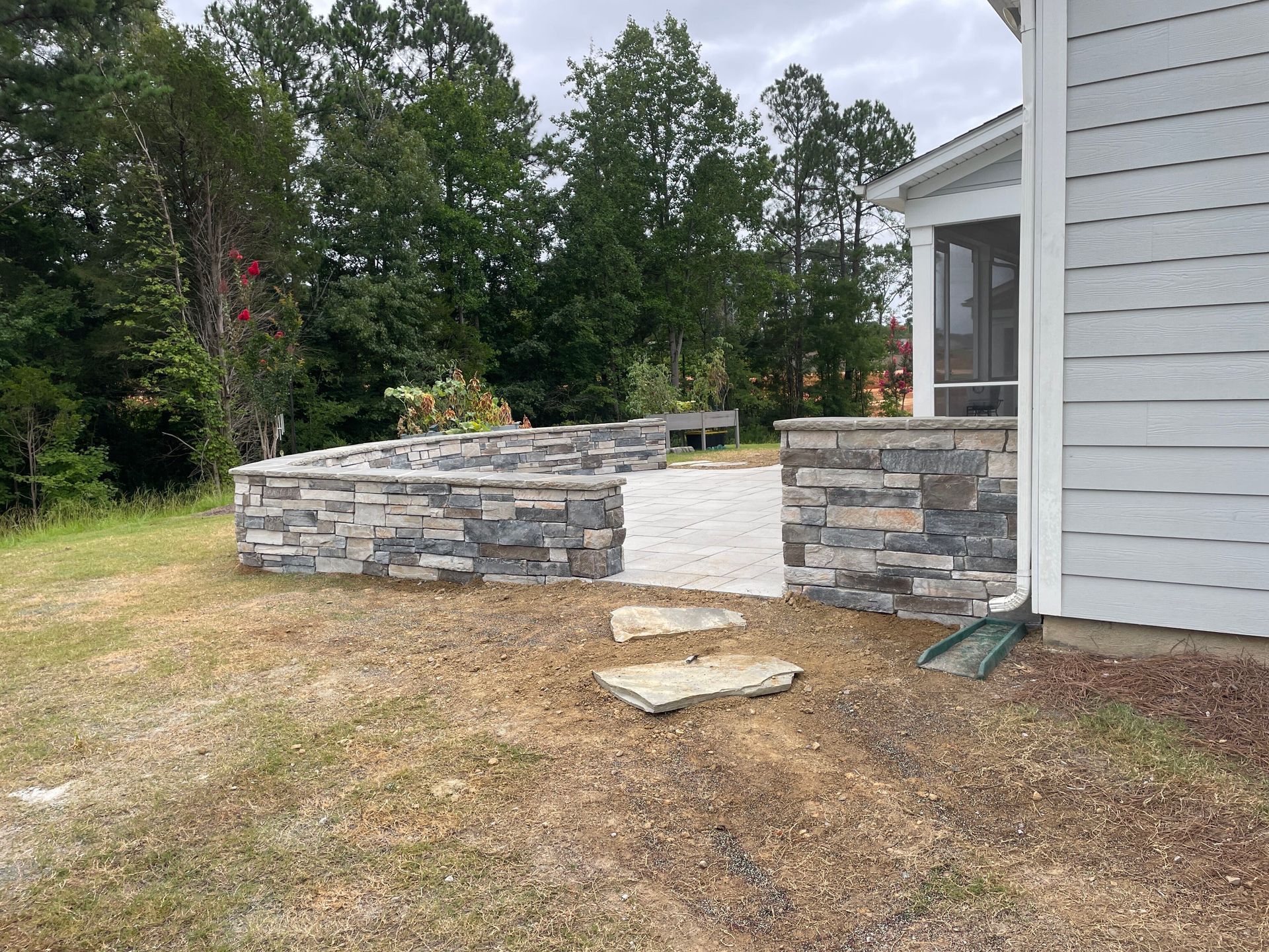 Stone retaining wall and steps adjacent to a light-colored house with a screened porch, set in a grassy yard.