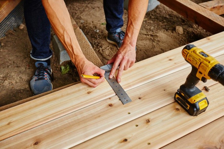 Hands measuring and marking a wooden board on a construction site with a drill nearby