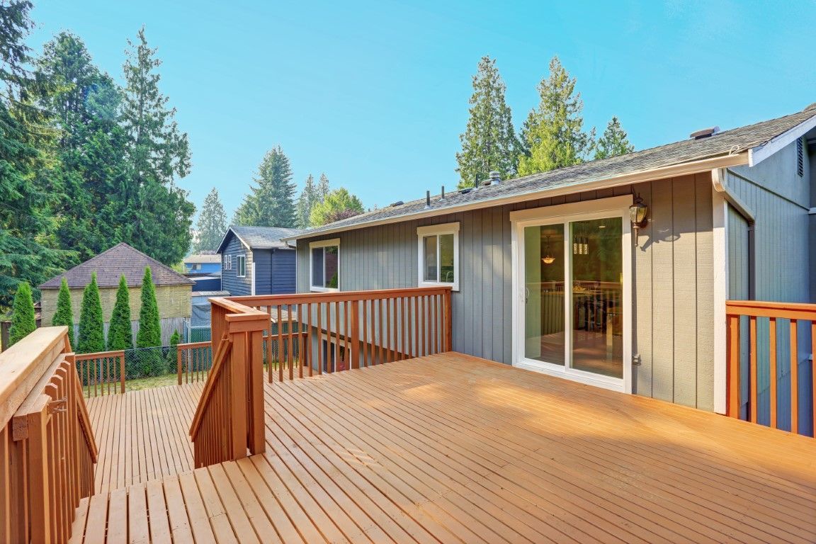 Wooden deck with railing beside a house, overlooking trees under a clear blue sky.