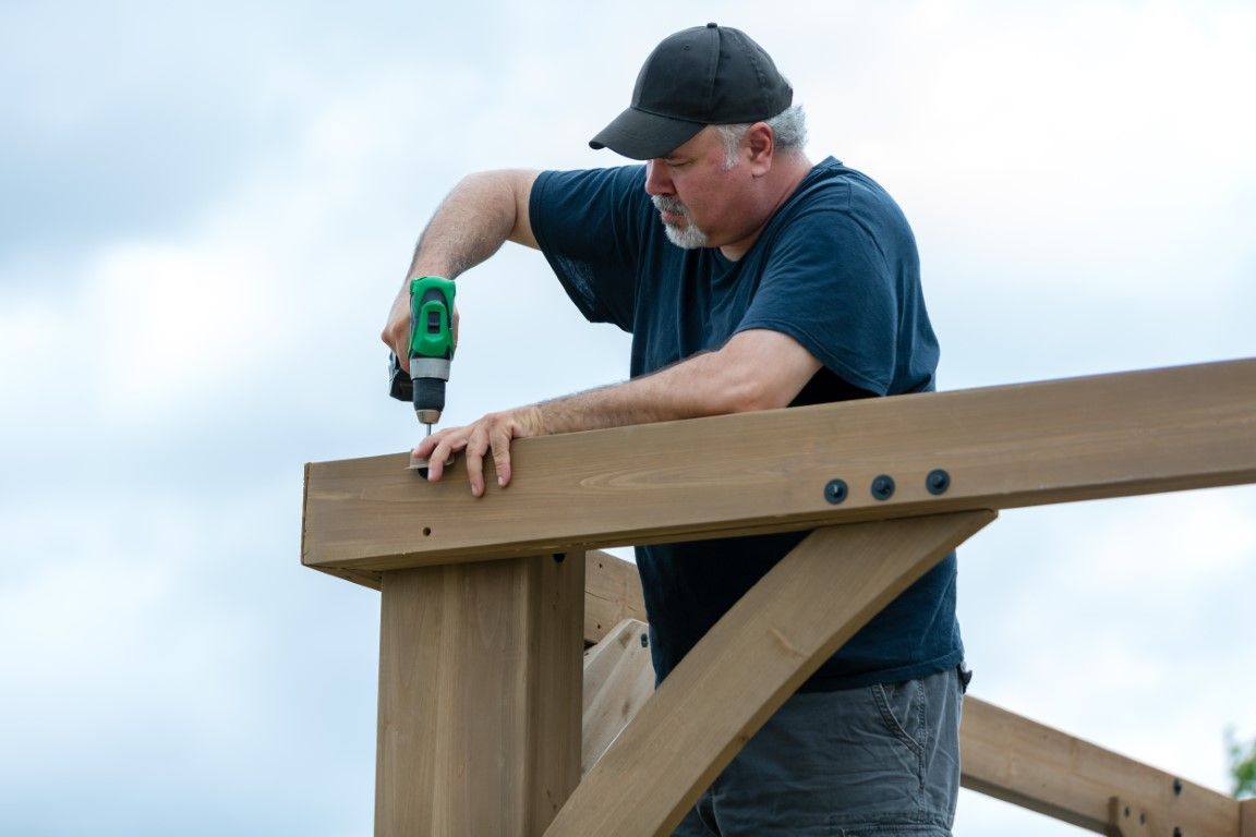 Man using a drill to fasten a wooden beam outdoors under a cloudy sky