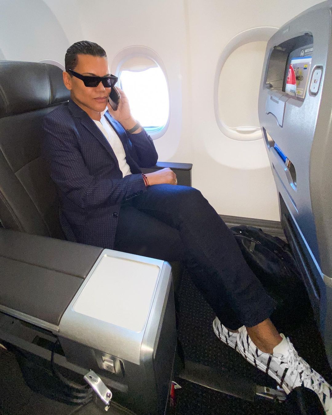 A man wearing sunglasses is sitting on an airplane talking on a cell phone