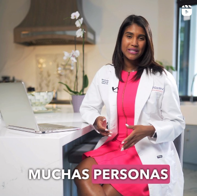 A woman in a pink dress and white coat is sitting at a counter with a laptop and the words muchas personas below her