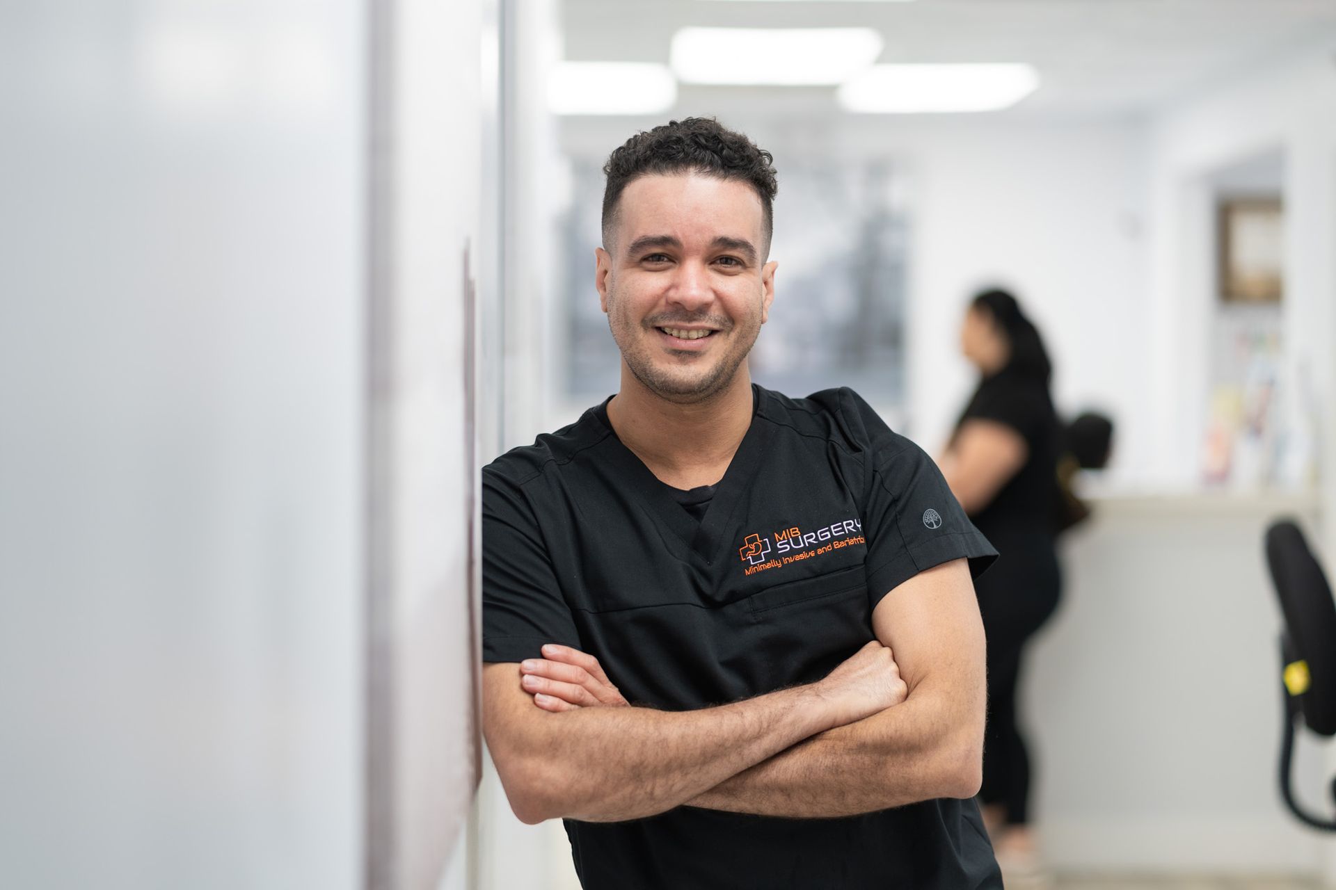 A man in a scrub is leaning against a wall with his arms crossed and smiling.