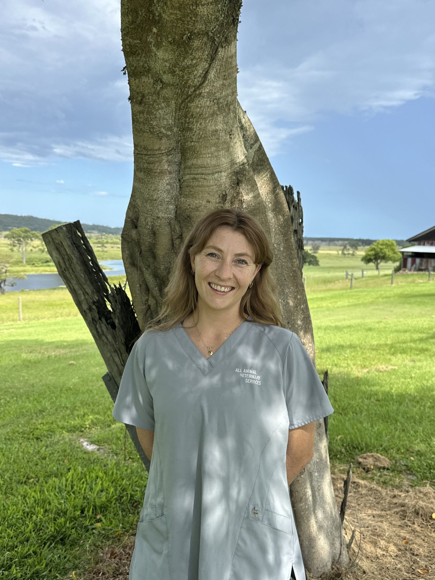 Woman in gray scrubs stands near a tree outdoors, smiling. Green field and cloudy sky background.