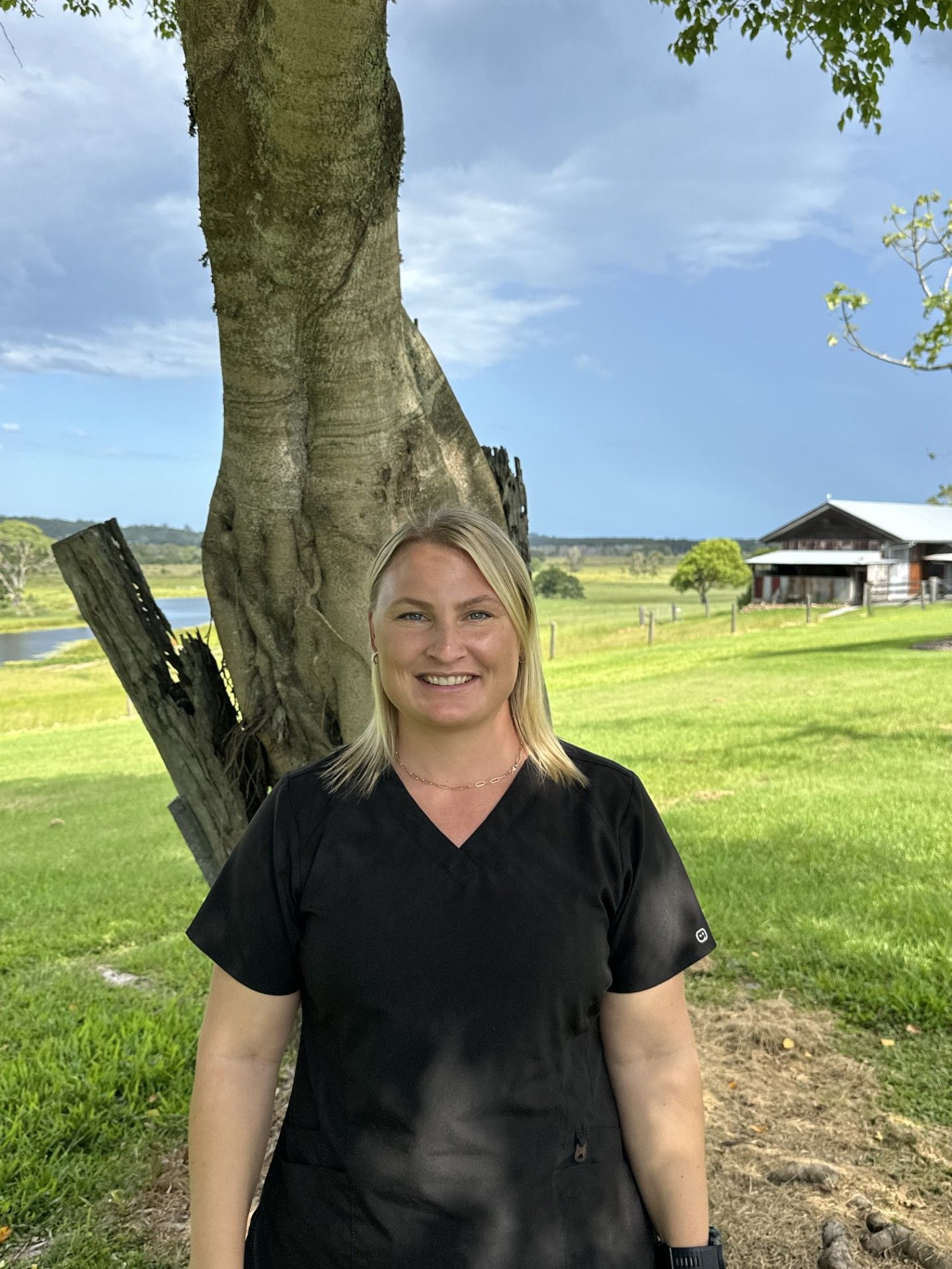 Woman in black scrubs smiles near a tree, with a barn and field in the background.