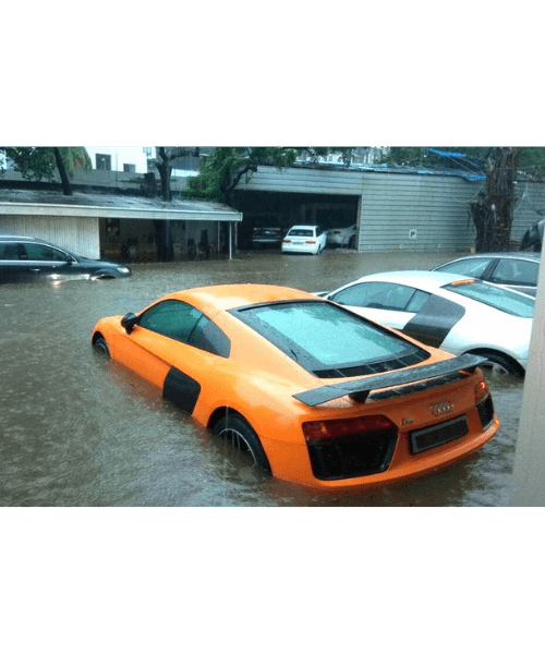 Um Audi R8 laranja, esportivo, submerso em água de enchente, estacionado próximo a outros veículos.