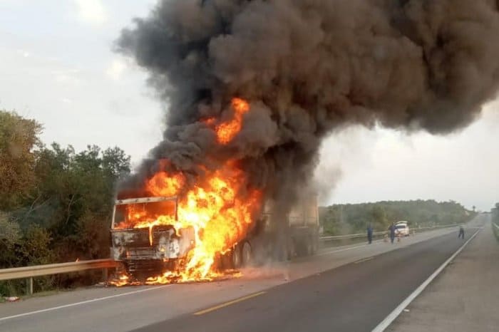 Caminhão em chamas na estrada, com labaredas e uma densa fumaça preta. Pessoas observam do local.