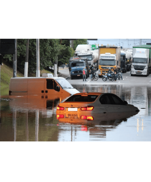Estrada alagada com carro e van parcialmente submersos, caminhões e veículos ao fundo. Água marrom, céu nublado.