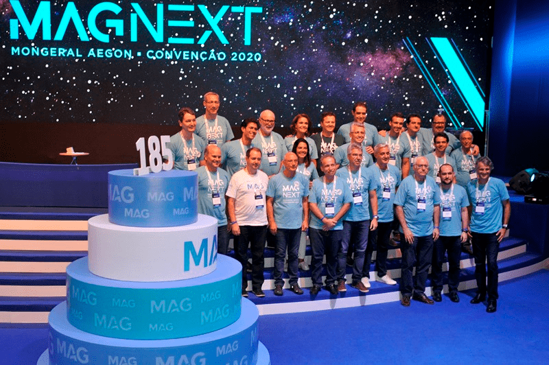 Um grupo de pessoas vestindo camisetas iguais posa com um bolo de vários andares em uma convenção da MAGNEXT.