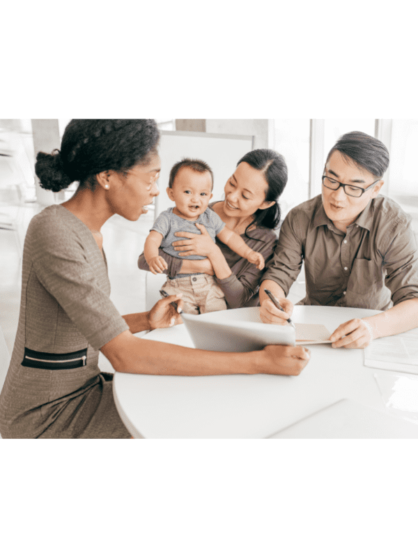 Uma mulher atende um casal que segura um bebê sentado à mesa. Eles revisam documentos e conversam.