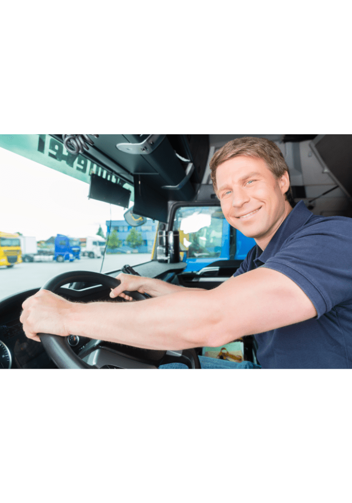 Motorista de caminhão sorridente na cabine de um caminhão semirreboque, com as mãos no volante. Dia ensolarado, com outros veículos ao fundo.