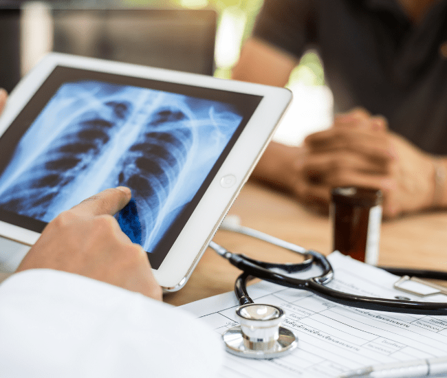 Médico apontando para uma radiografia de tórax em um tablet, paciente sentado próximo, estetoscópio e papéis sobre a mesa.