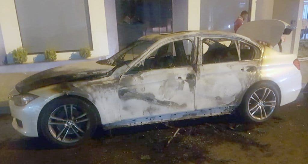 Carro branco incendiado, provavelmente um BMW, com o capô aberto, danificado em frente a um prédio.