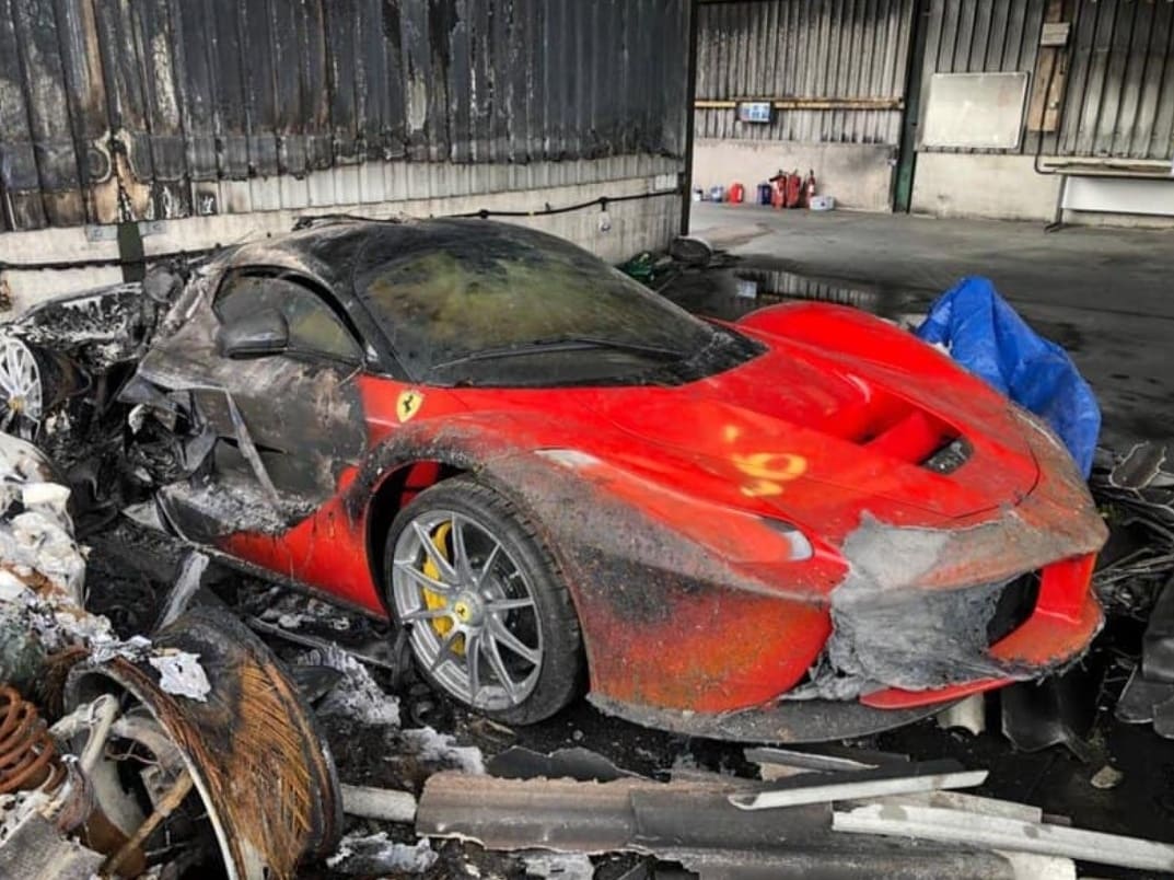 Ferrari vermelha queimada dentro de um prédio danificado, coberta de fuligem e destroços.
