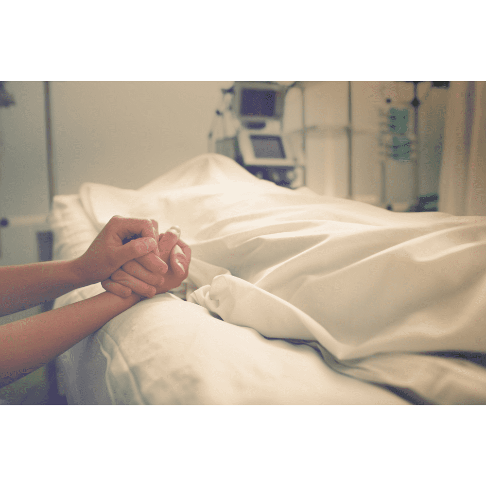 Mãos entrelaçadas em sinal de conforto ao lado de uma cama de hospital coberta com um lençol branco. Equipamentos médicos ao fundo.