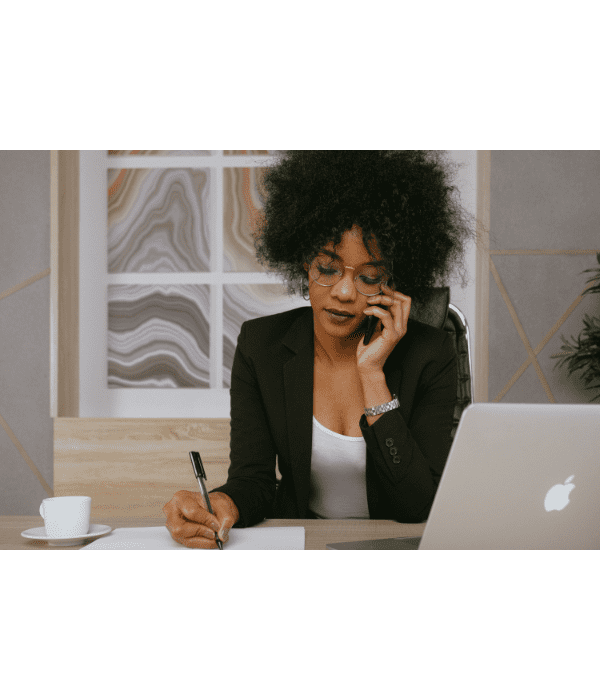 Mulher de blazer ao telefone, escrevendo em uma mesa com laptop e xícara de café.