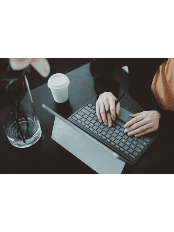 Pessoa digitando em um teclado de laptop preto, com uma xícara de café e um vaso sobre uma mesa escura.