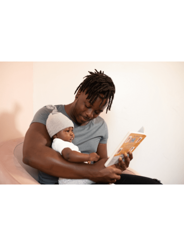 Homem lendo um livro para um bebê que usa chapéu. Eles estão sentados juntos em um sofá rosa.