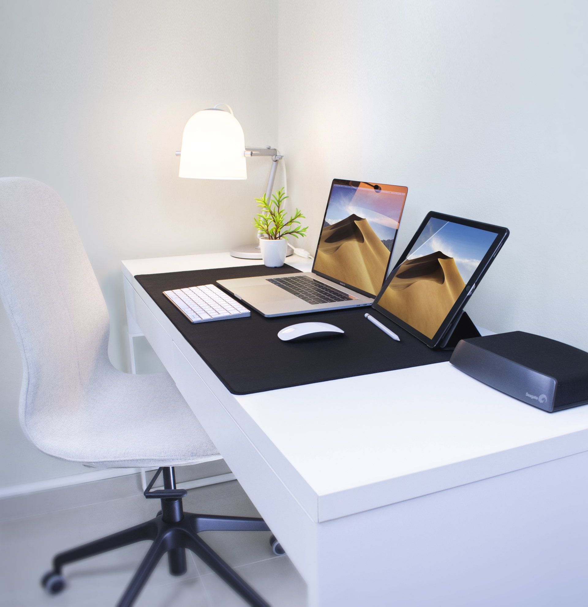 Mesa branca com laptops, tablet, mouse, teclado e luminária. Uma cadeira está à esquerda da mesa.