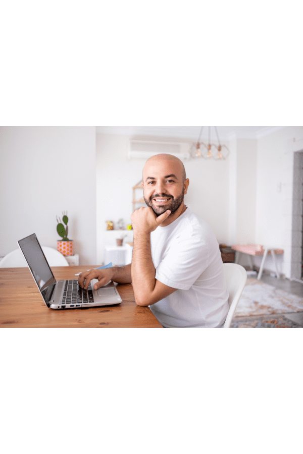 Homem careca de camisa branca, sorrindo, sentado a uma mesa de madeira com um laptop. Ambiente interno com luz natural.