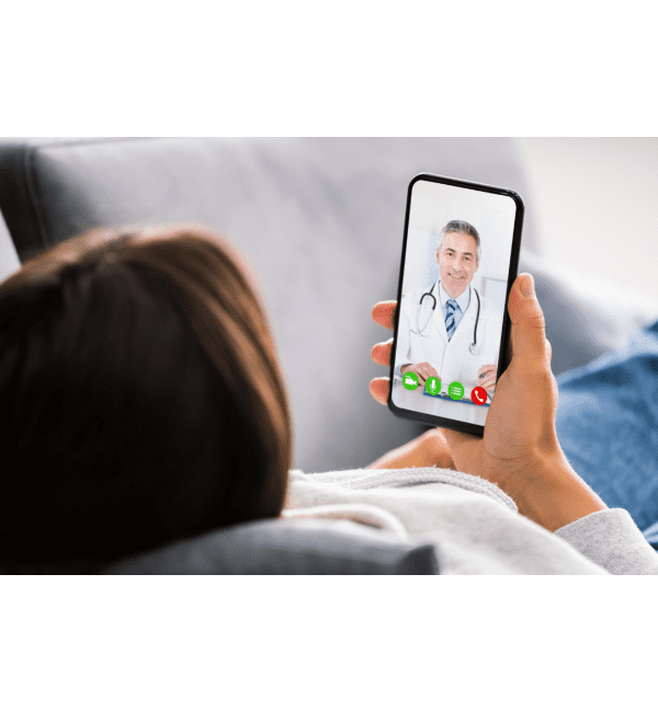 Uma pessoa está reclinada em um sofá, segurando um smartphone durante uma videochamada de telemedicina com um médico sorridente.