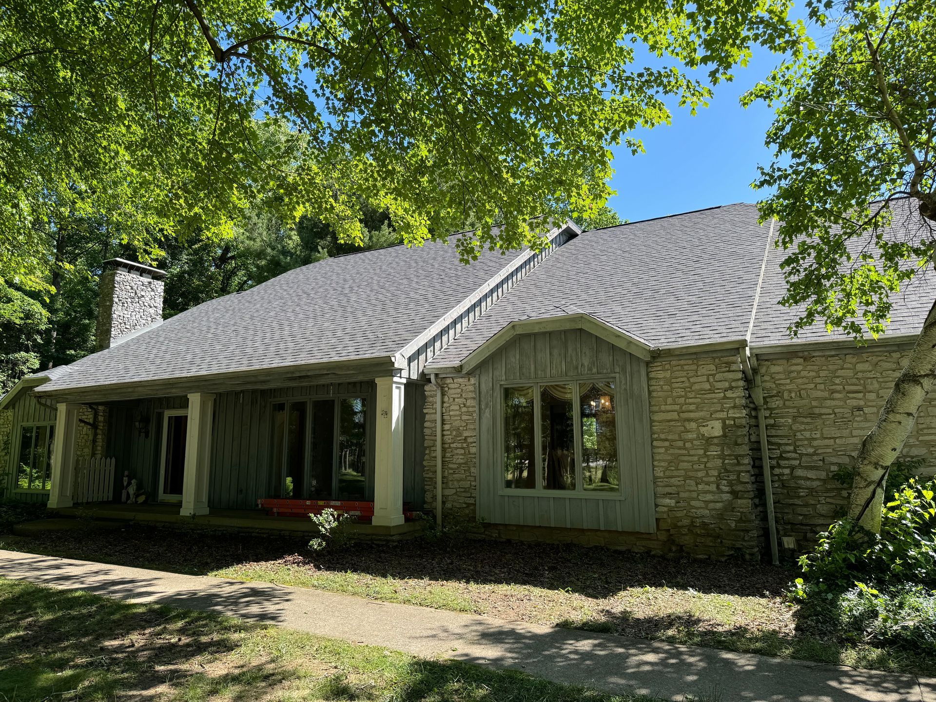 A large stone house with a gray roof and a porch surrounded by trees.