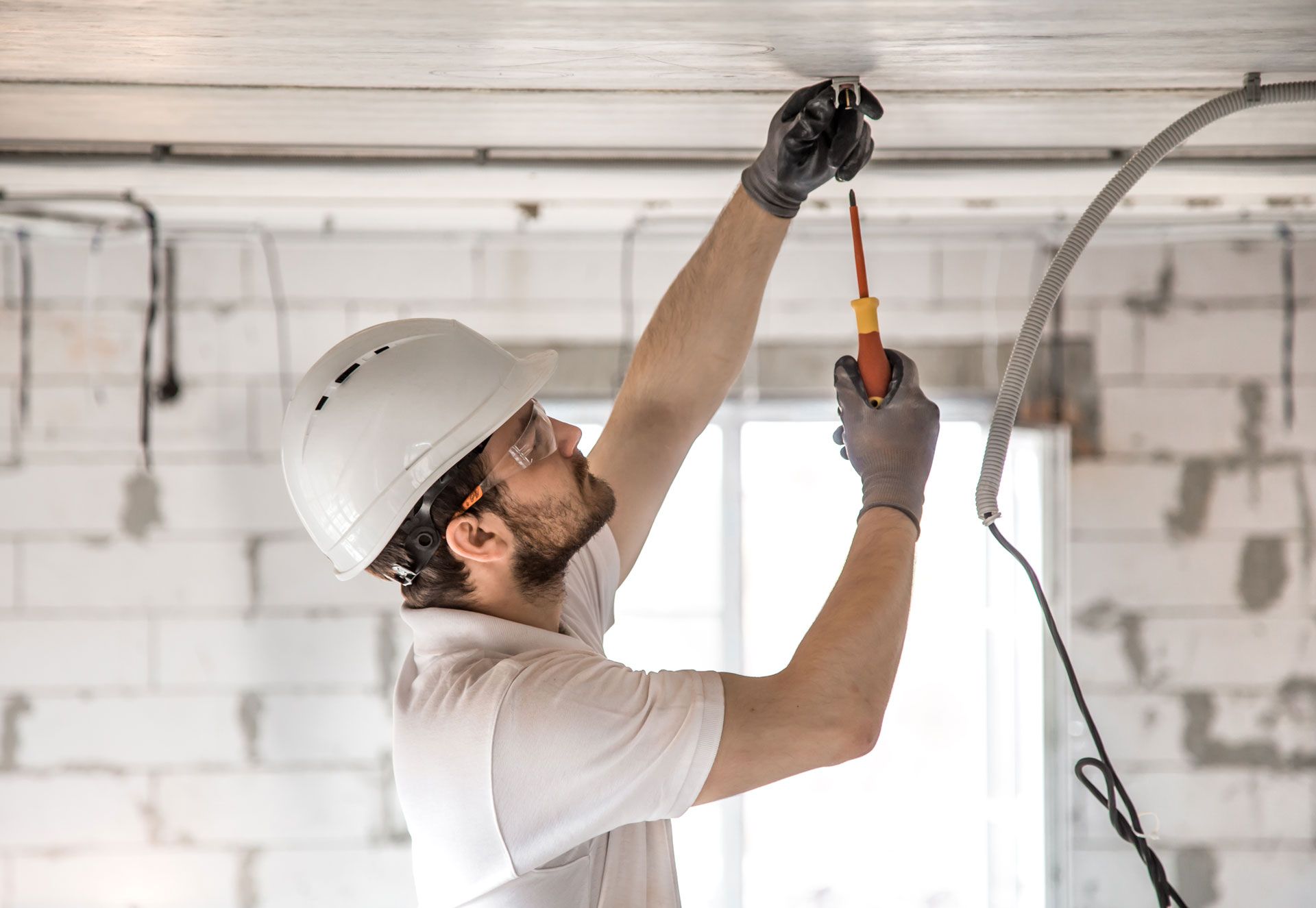 Worker in a hard hat fixing ceiling wiring with pliers in an unfinished room