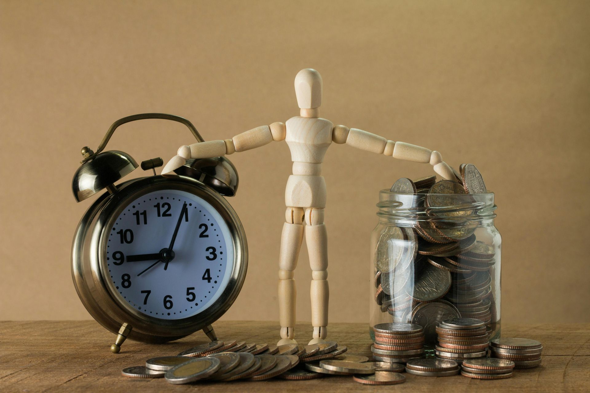 Wooden mannequin with arms outstretched, standing between a brass alarm clock and a jar overflowing with coins.  Setting: wooden table against a neutral background.