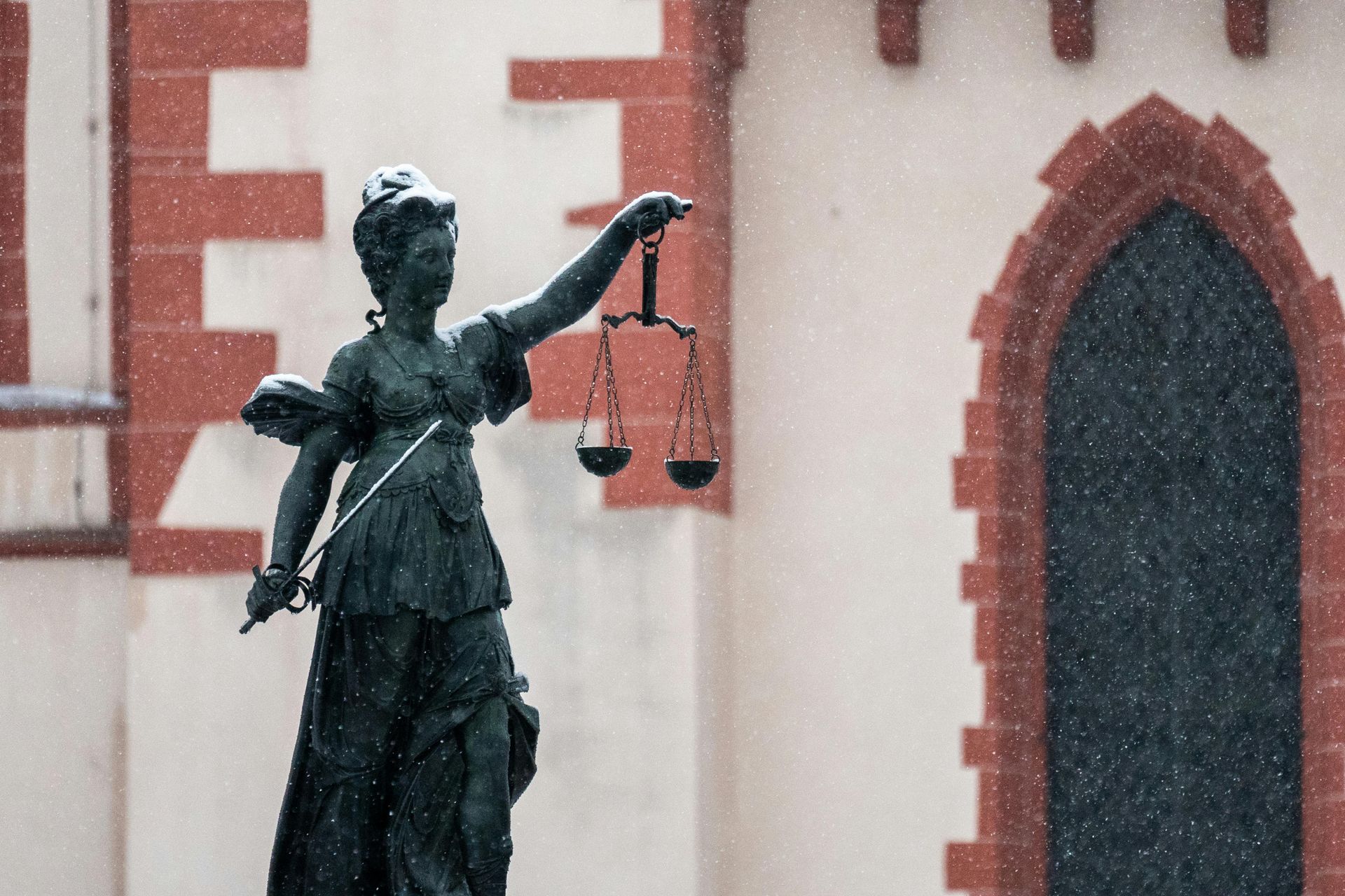 A bronze statue of Lady Justice holding scales and a sword stands against a red-trimmed stone building during a snowfall.