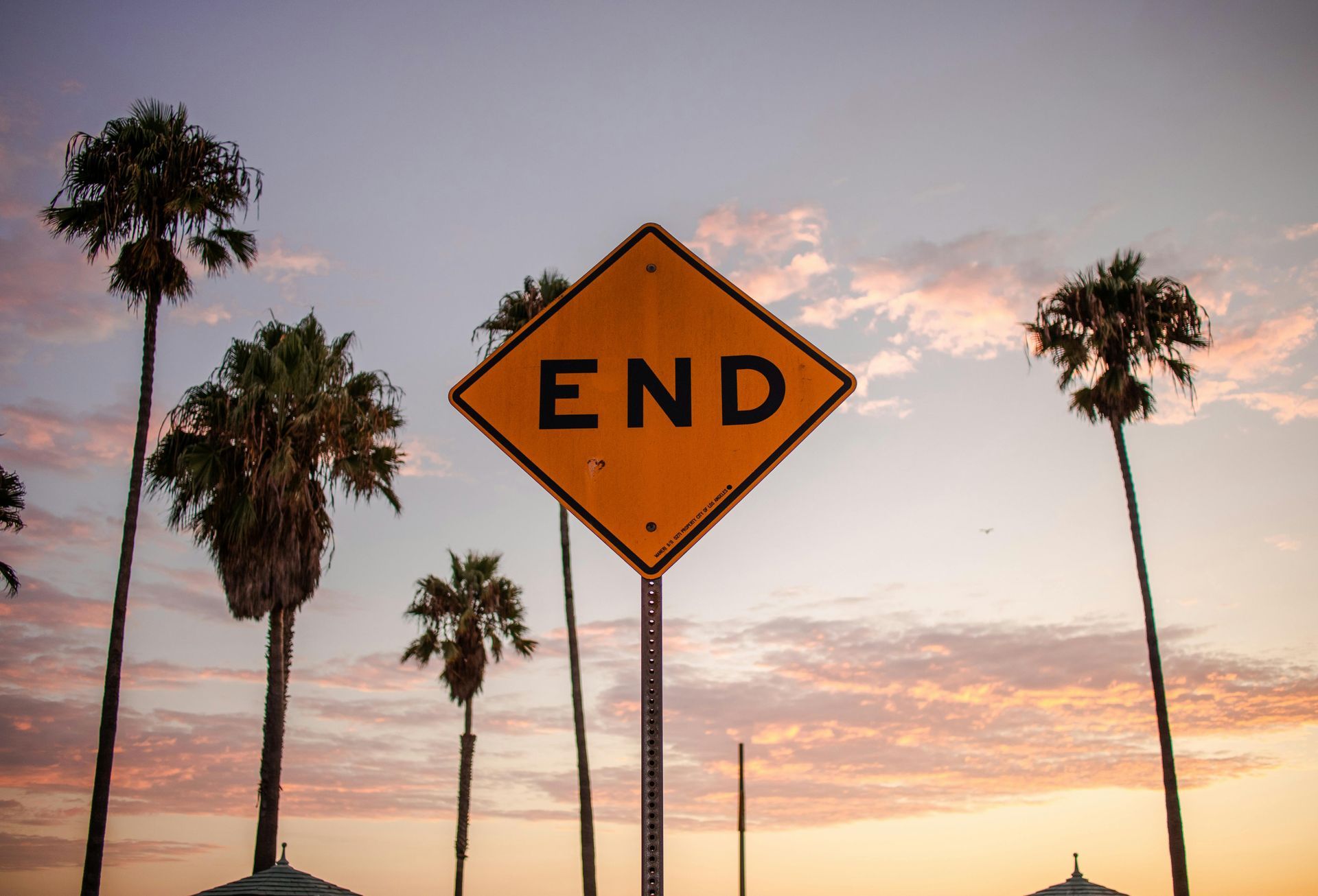 Yellow END road sign in front of palm trees against a sunset sky.