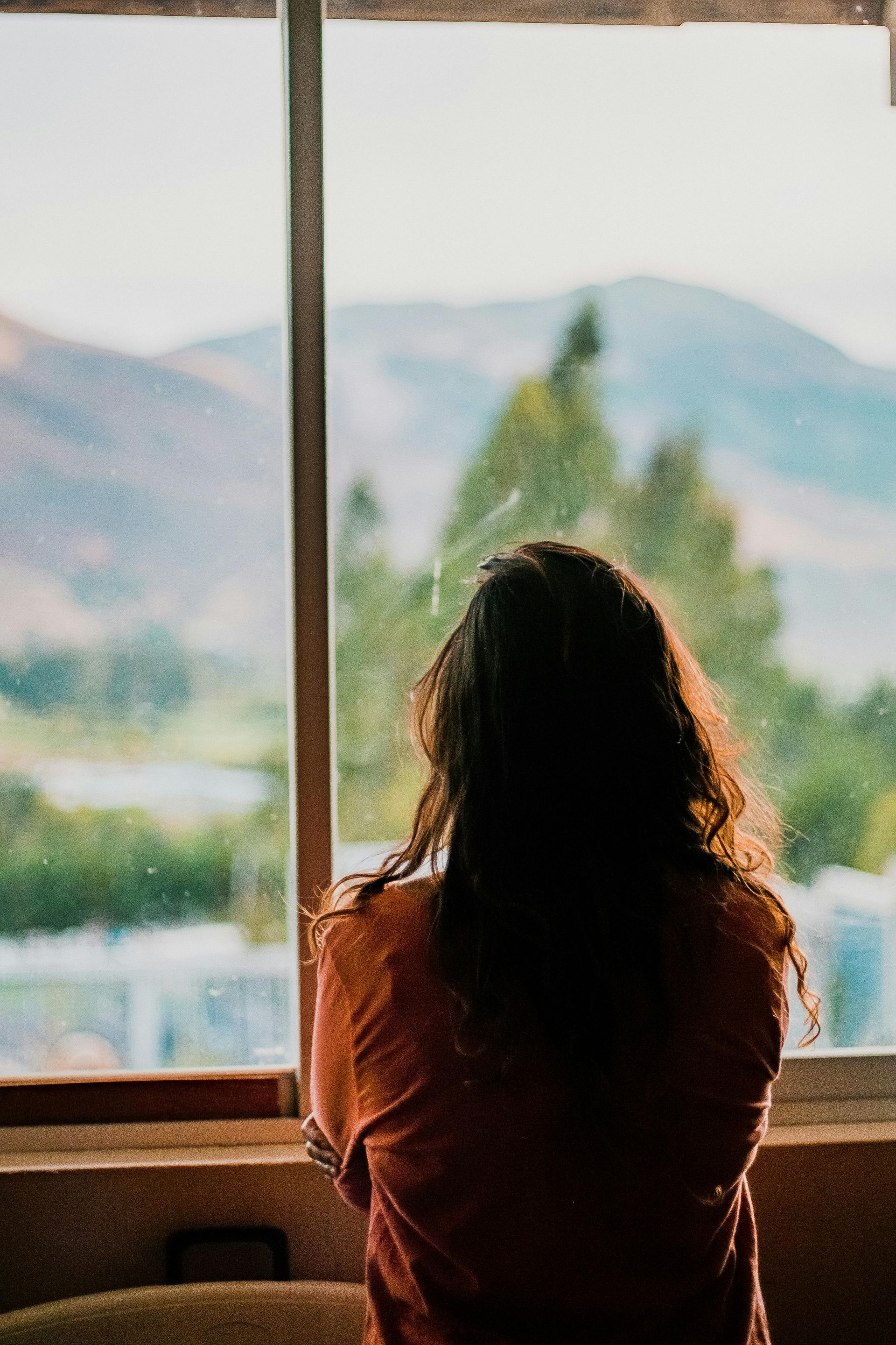 Person gazing out a window at a mountain view. Backlit, long wavy hair, orange shirt, blurred background. Waiting.